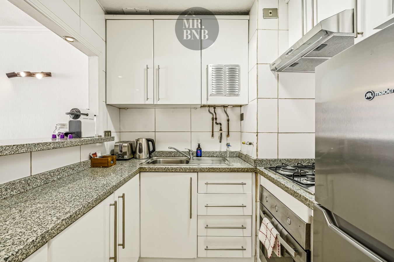 Cocina en L con muebles blancos, campana extractora, cocina a gas y refrigerador