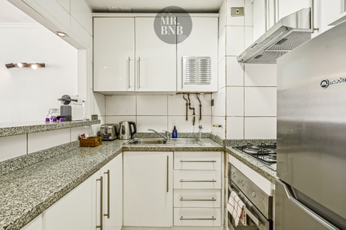 Cocina en L con muebles blancos, campana extractora, cocina a gas y refrigerador