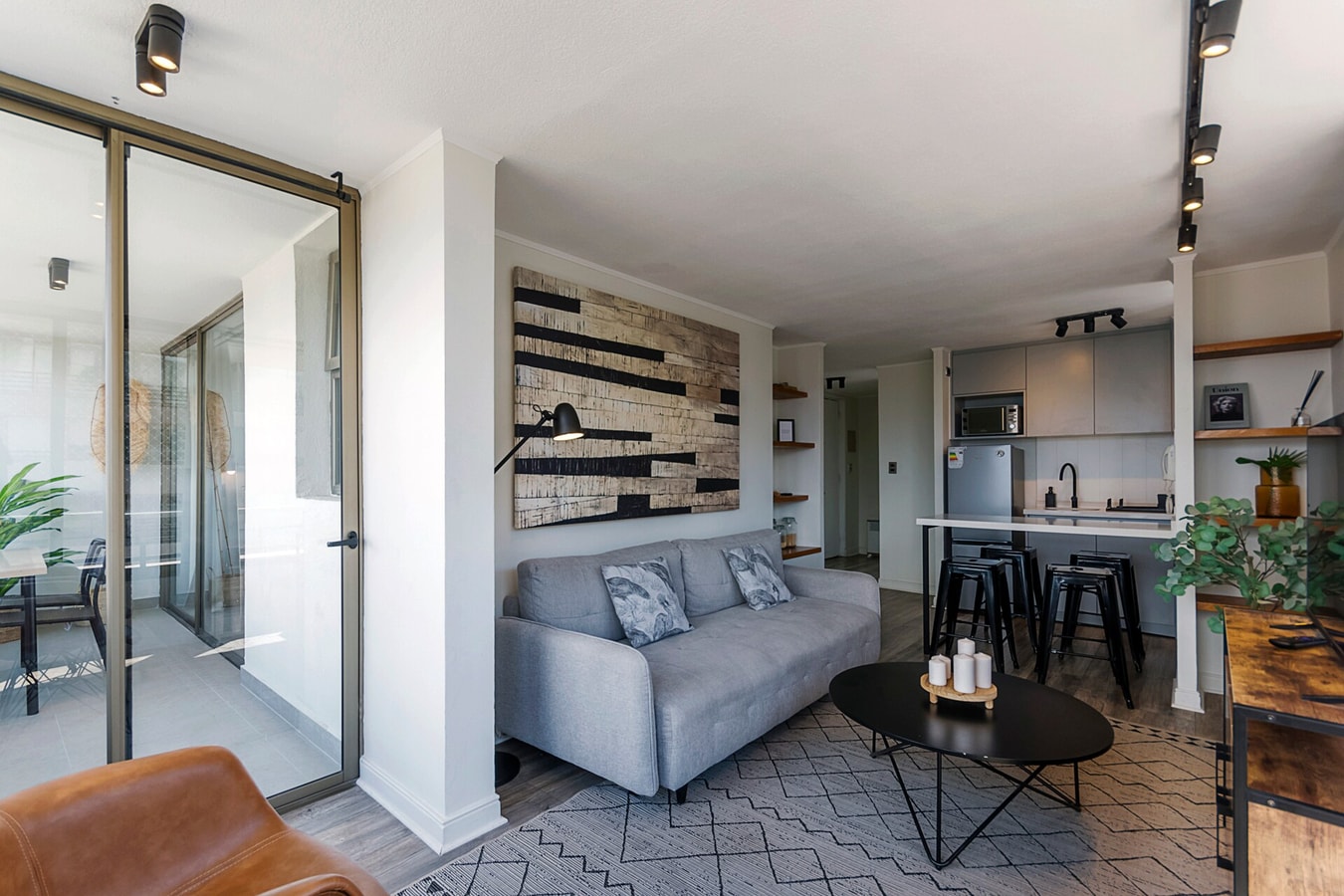 Living con sofá gris, mesa de centro, sillón de cuero y cocina al fondo con barra y taburetes