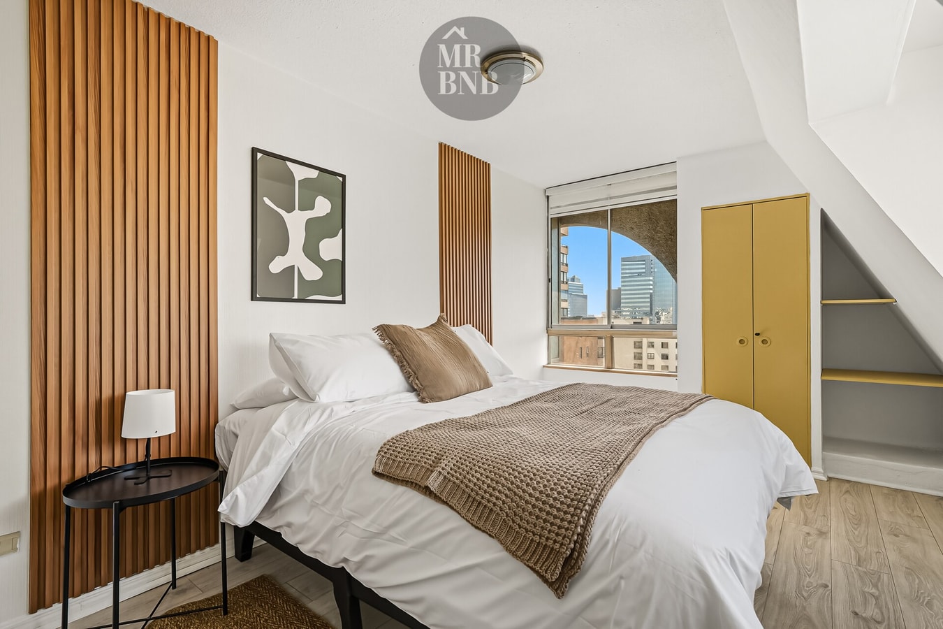 Dormitorio con panel de madera, cama con manta y ventana con vista a la ciudad
