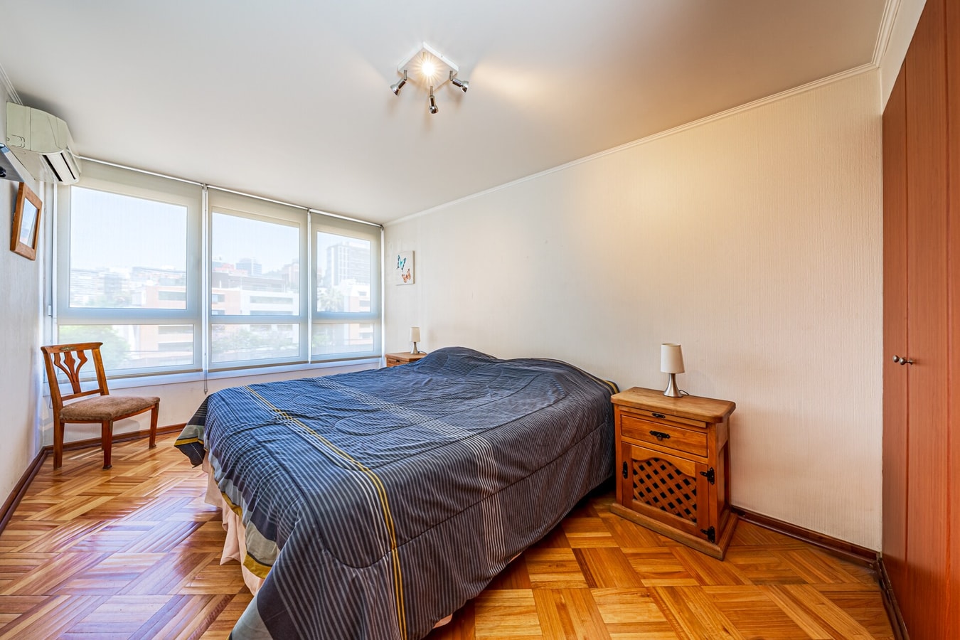 Dormitorio con cama, ventana amplia con vista a la ciudad, silla y equipo de aire acondicionado en el muro.