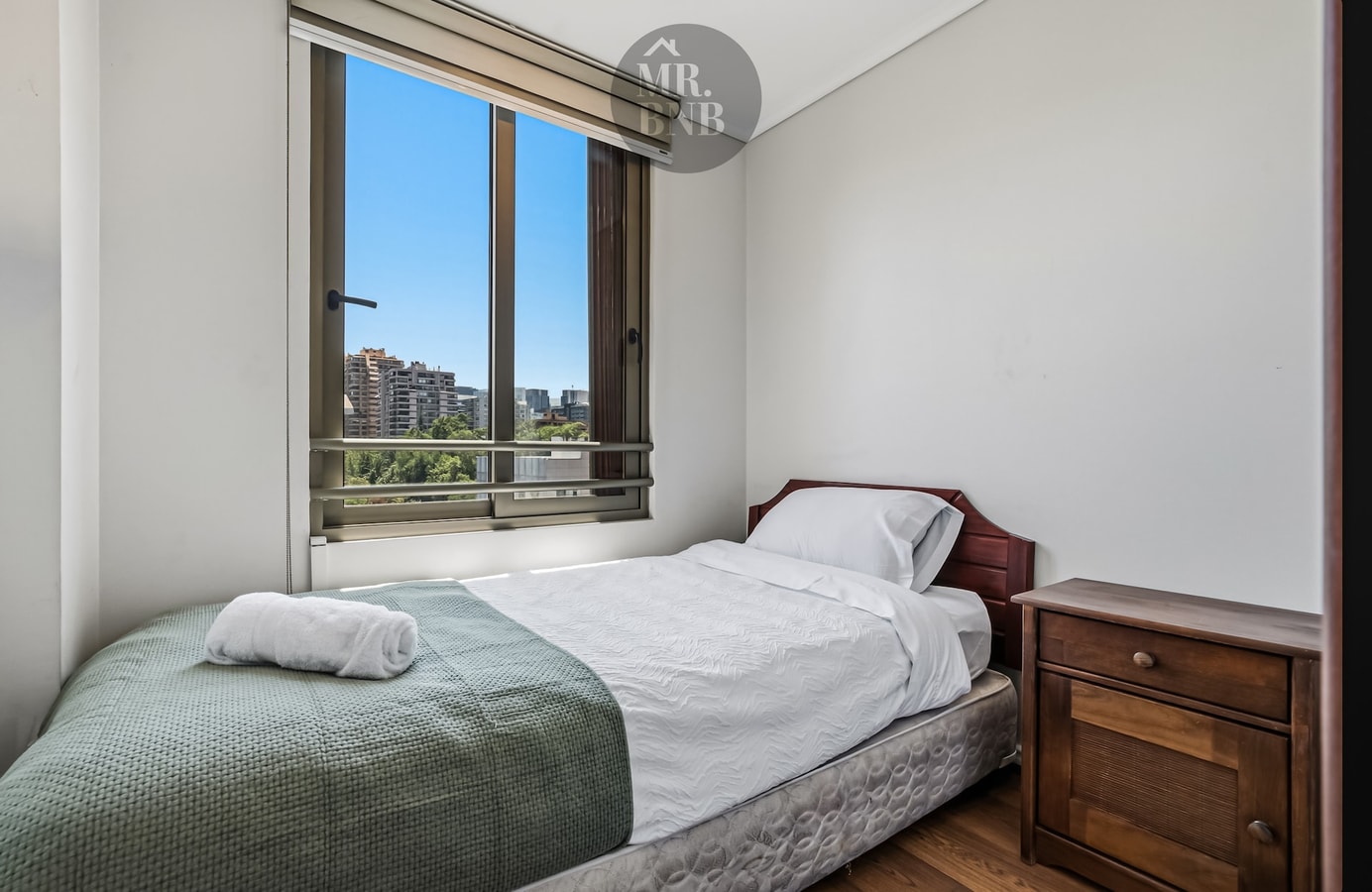 Dormitorio con cama individual, velador de madera y ventana con vista a edificios.