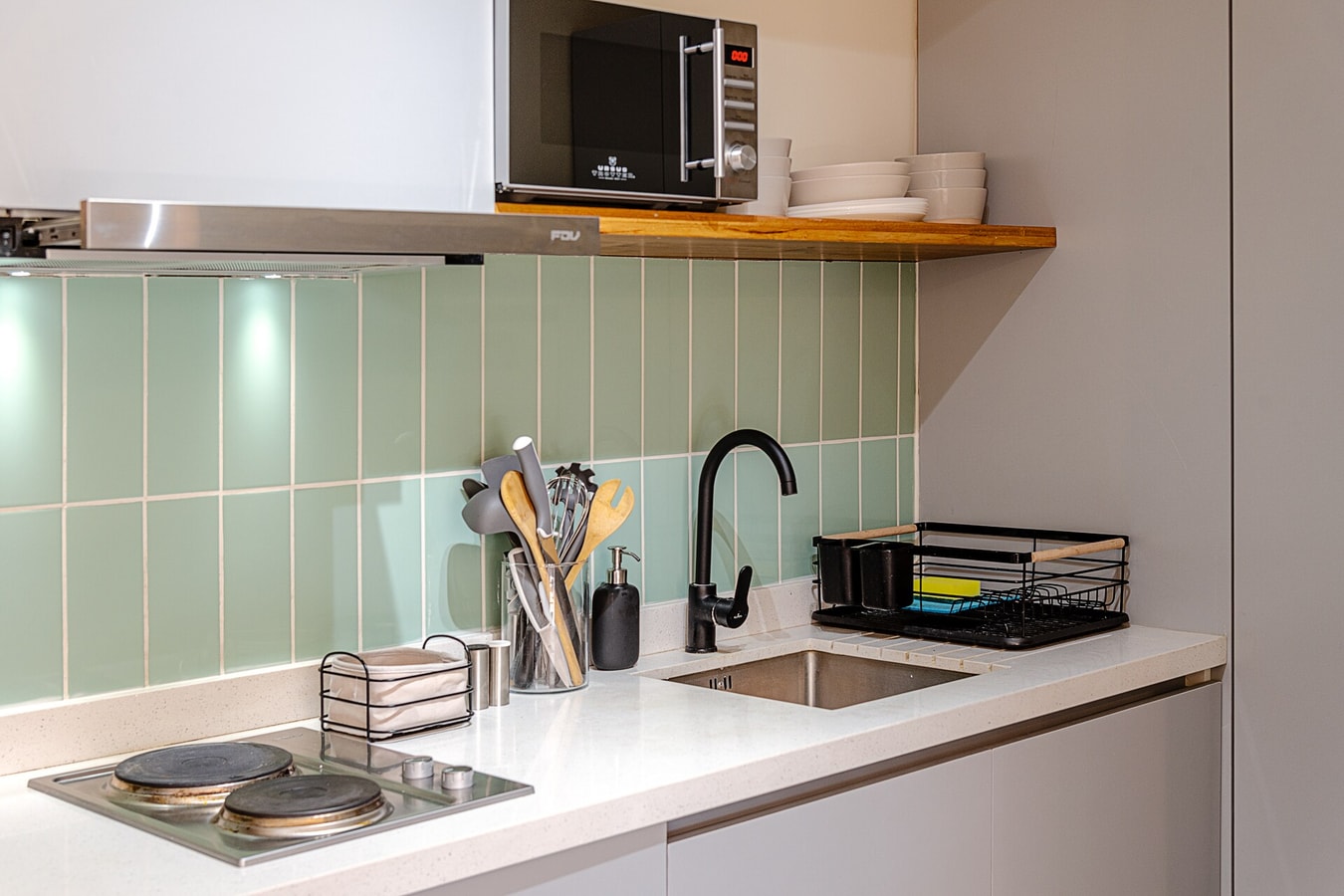 Detalle del mesón de cocina con encimera, campana, microondas, lavaplatos y utensilios sobre el azulejo verde agua