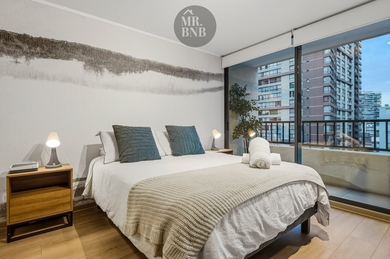 Dormitorio principal con cama de dos plazas, veladores con lámparas y ventanal con vista a la ciudad
