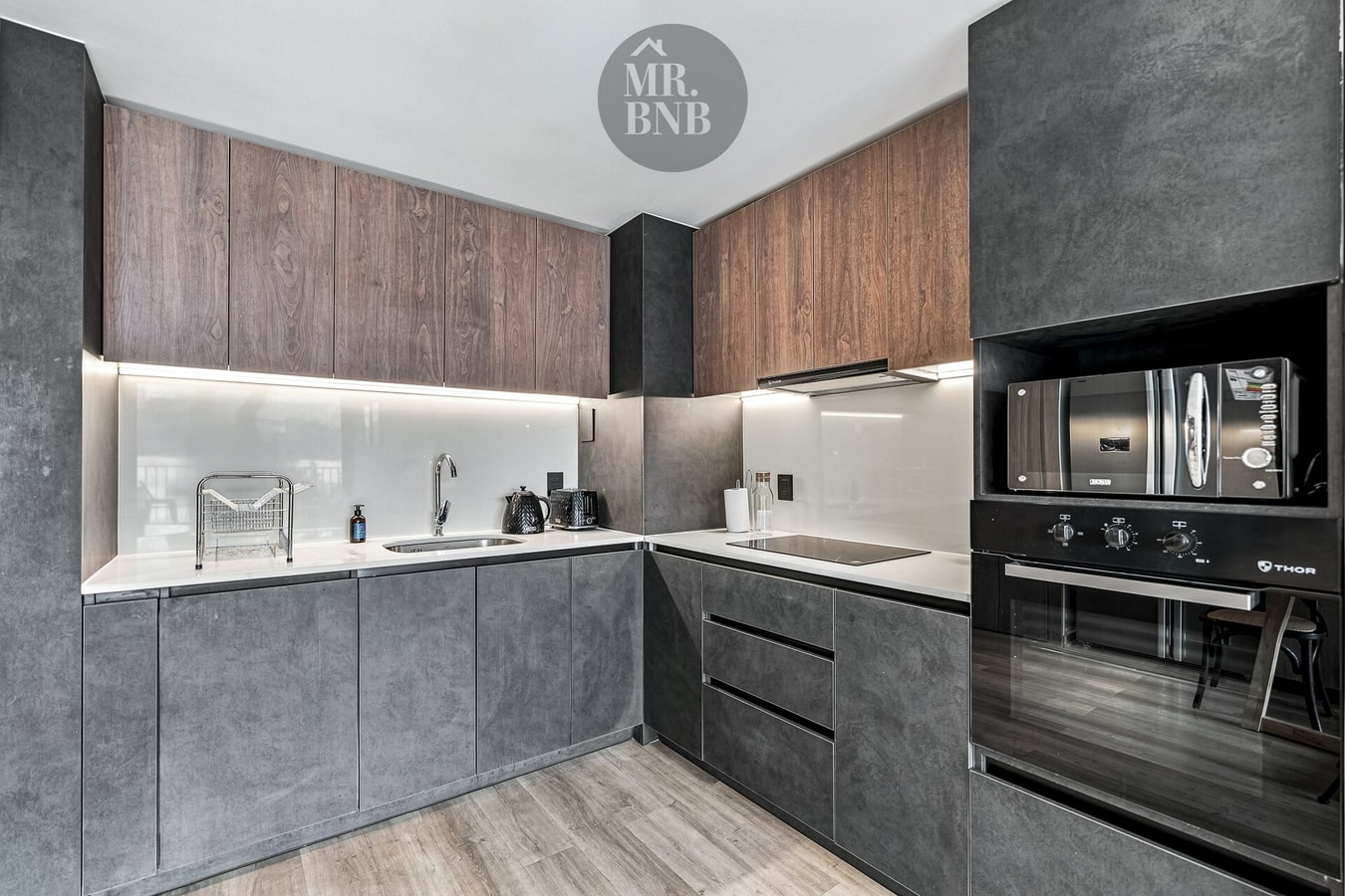 Cocina equipada con encimera eléctrica, horno, microondas y muebles en tonos madera y gris oscuro