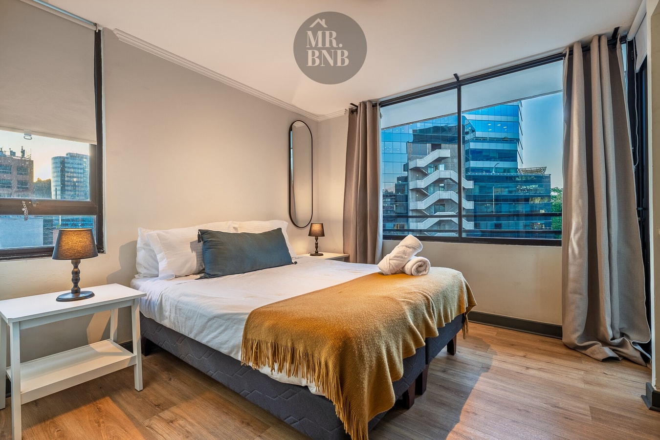 Dormitorio con cama de dos plazas, cubrecama azul y mostaza, y ventana con vista a edificios