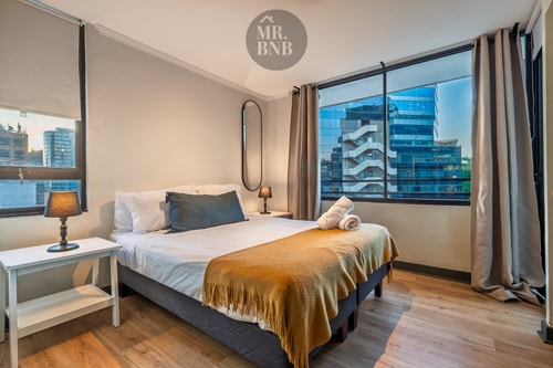 Dormitorio con cama de dos plazas, cubrecama azul y mostaza, y ventana con vista a edificios