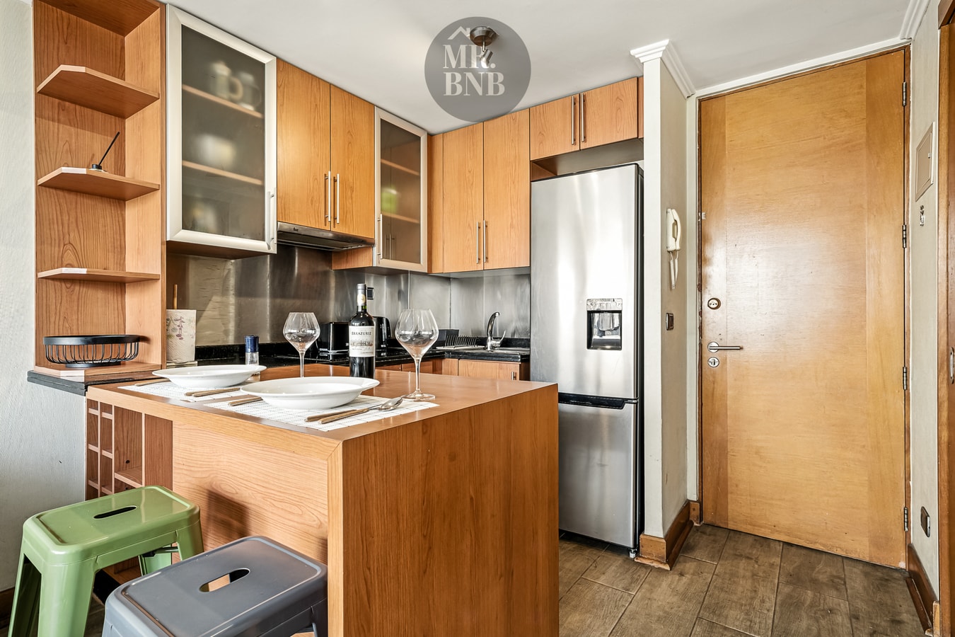 Cocina con isla, mesa puesta con vajilla y copas, taburetes, refrigerador y puerta de entrada