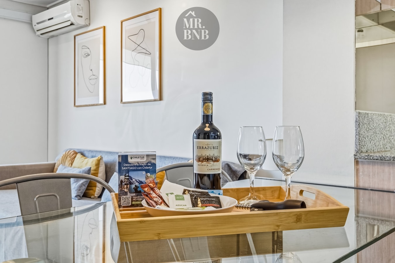 Bandeja de bienvenida con vino y copas sobre mesa de living, junto a sofá y cuadros en la pared.
