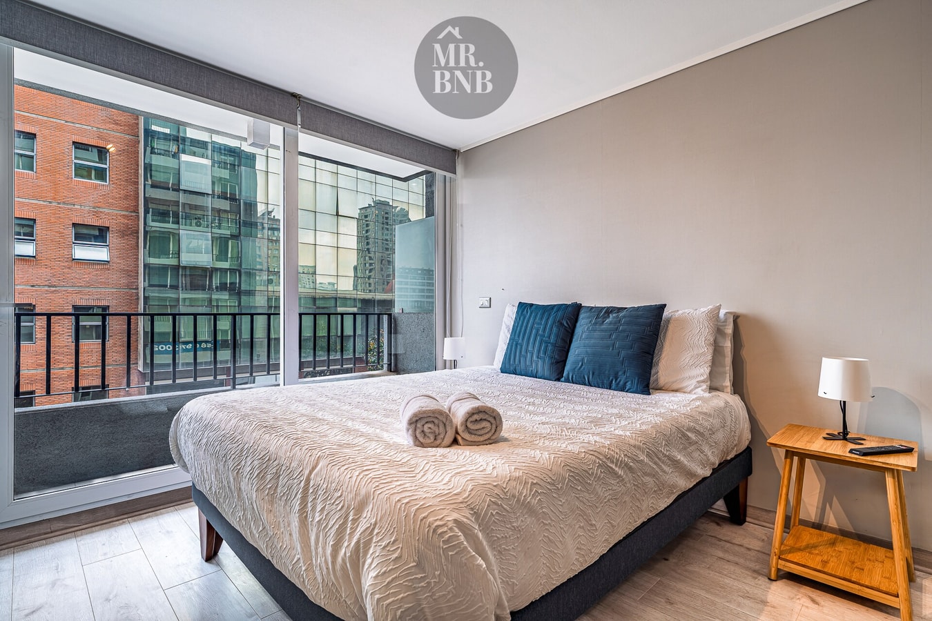 Dormitorio con cama de dos plazas, cojines azules, velador de madera y ventanal.