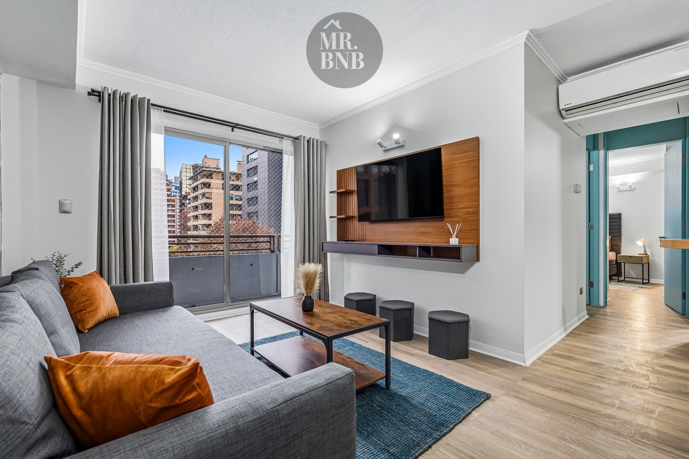 Living con sofá, mueble de TV, ventanal con vista a edificios y aire acondicionado