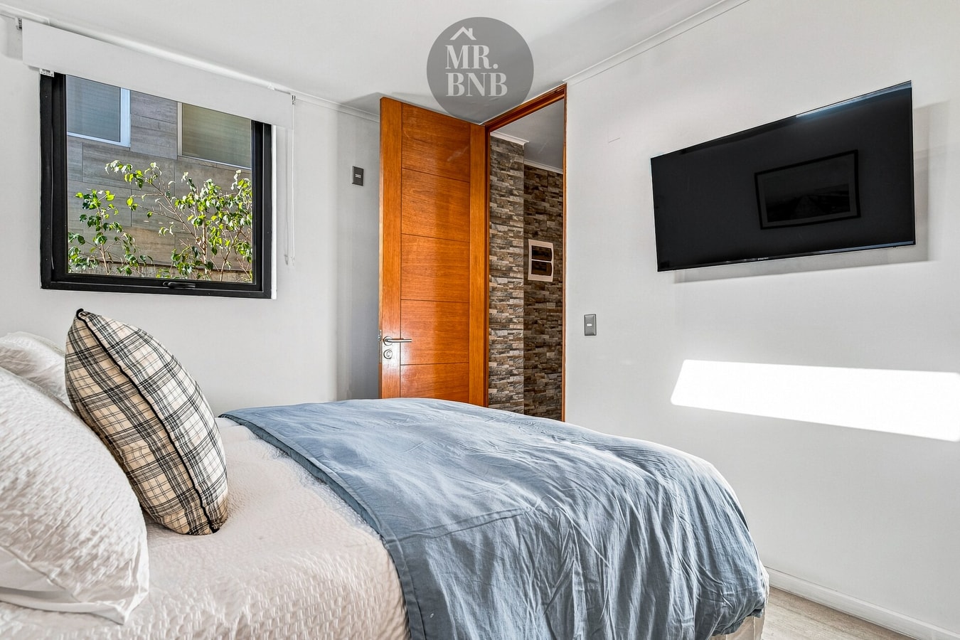 Dormitorio con cama, cubrecama celeste, TV en la pared y puerta de madera con textura