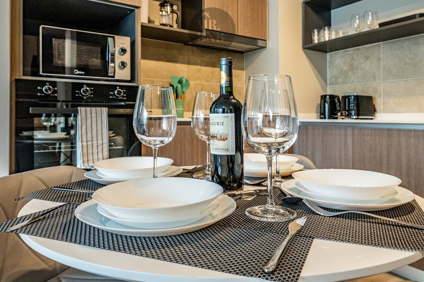 Mesa de comedor puesta con vajilla, copas de vino y microondas al fondo.