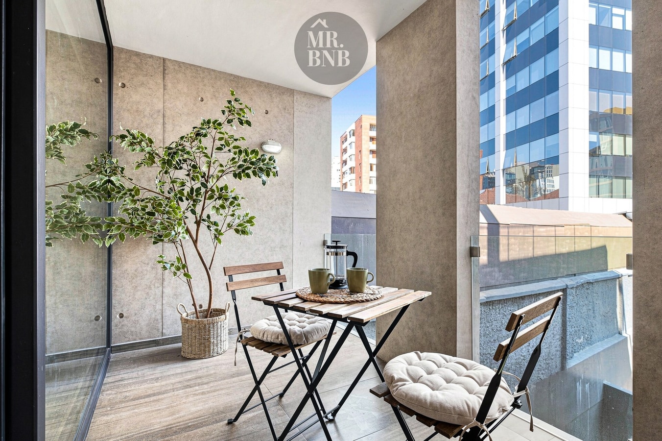 Terraza privada con mesa pequeña, sillas y planta, vista a edificios