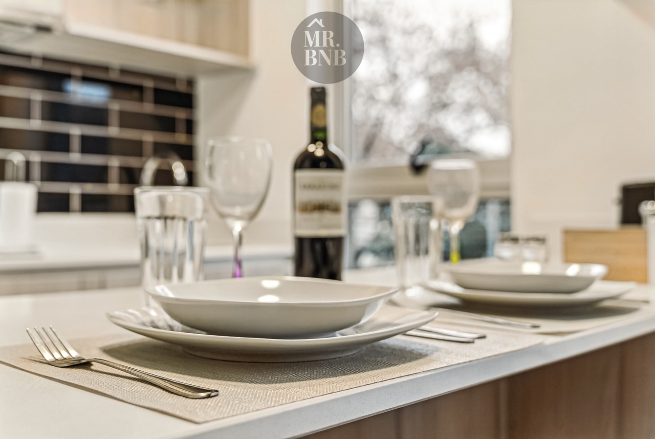 Detalle de mesa puesta sobre mesón de cocina, con copas, platos y una botella de vino