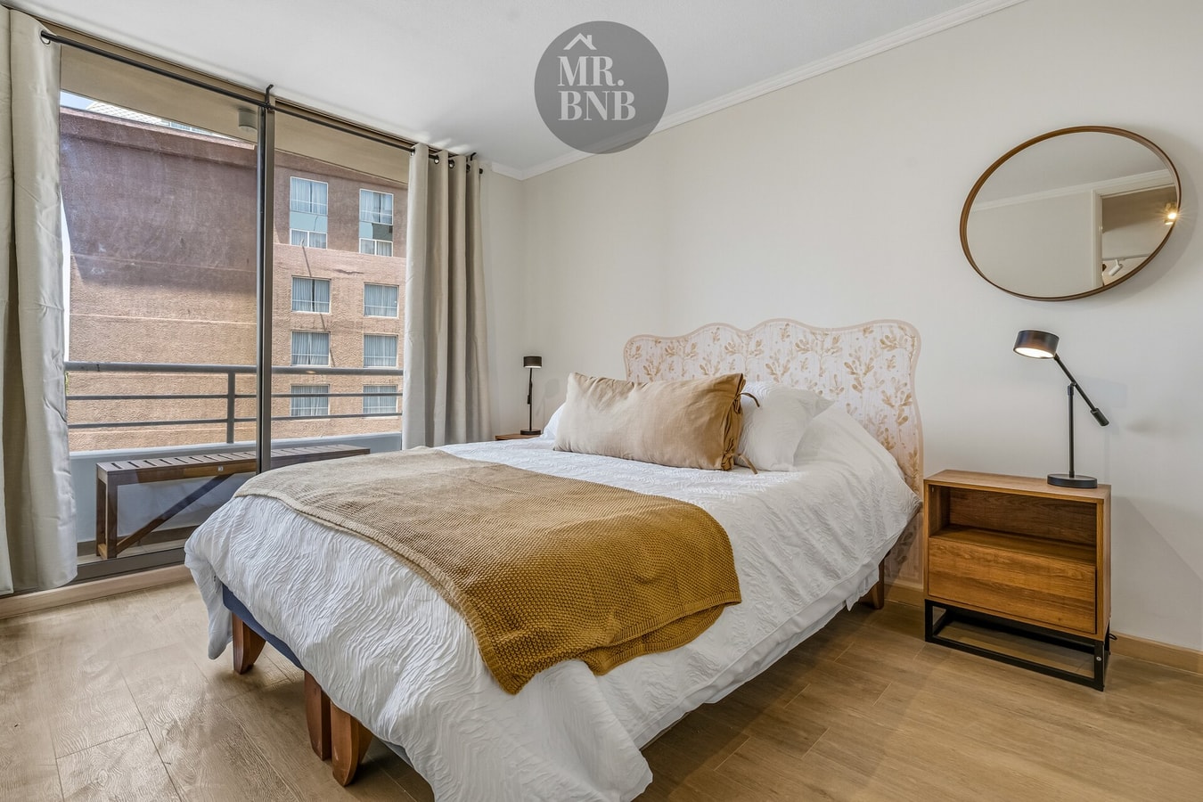 Dormitorio con cama, manta dorada, espejo redondo y velador con lámpara