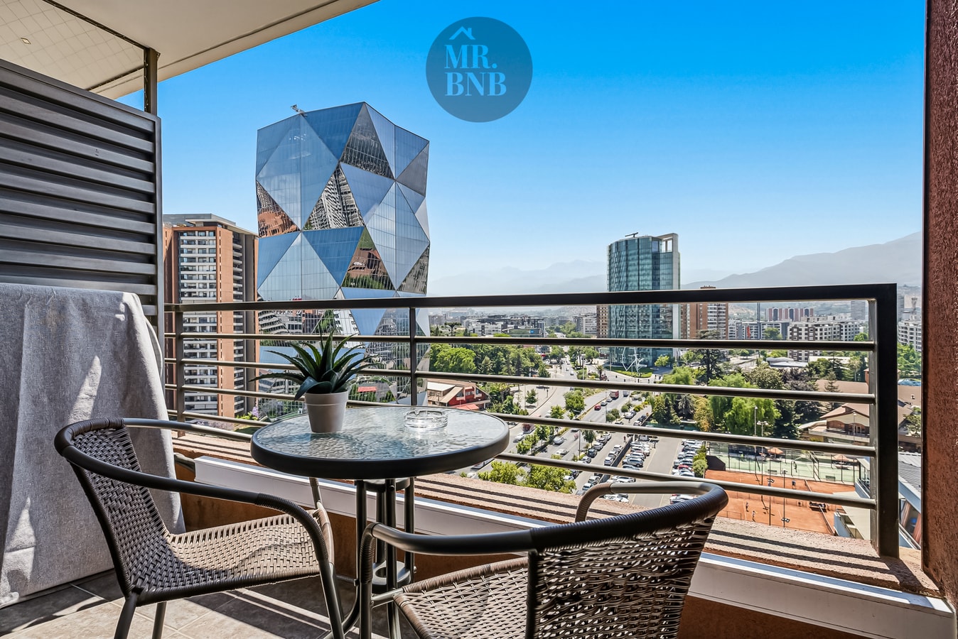 Terraza privada con mesa y dos sillas, vista a edificios y cordillera.