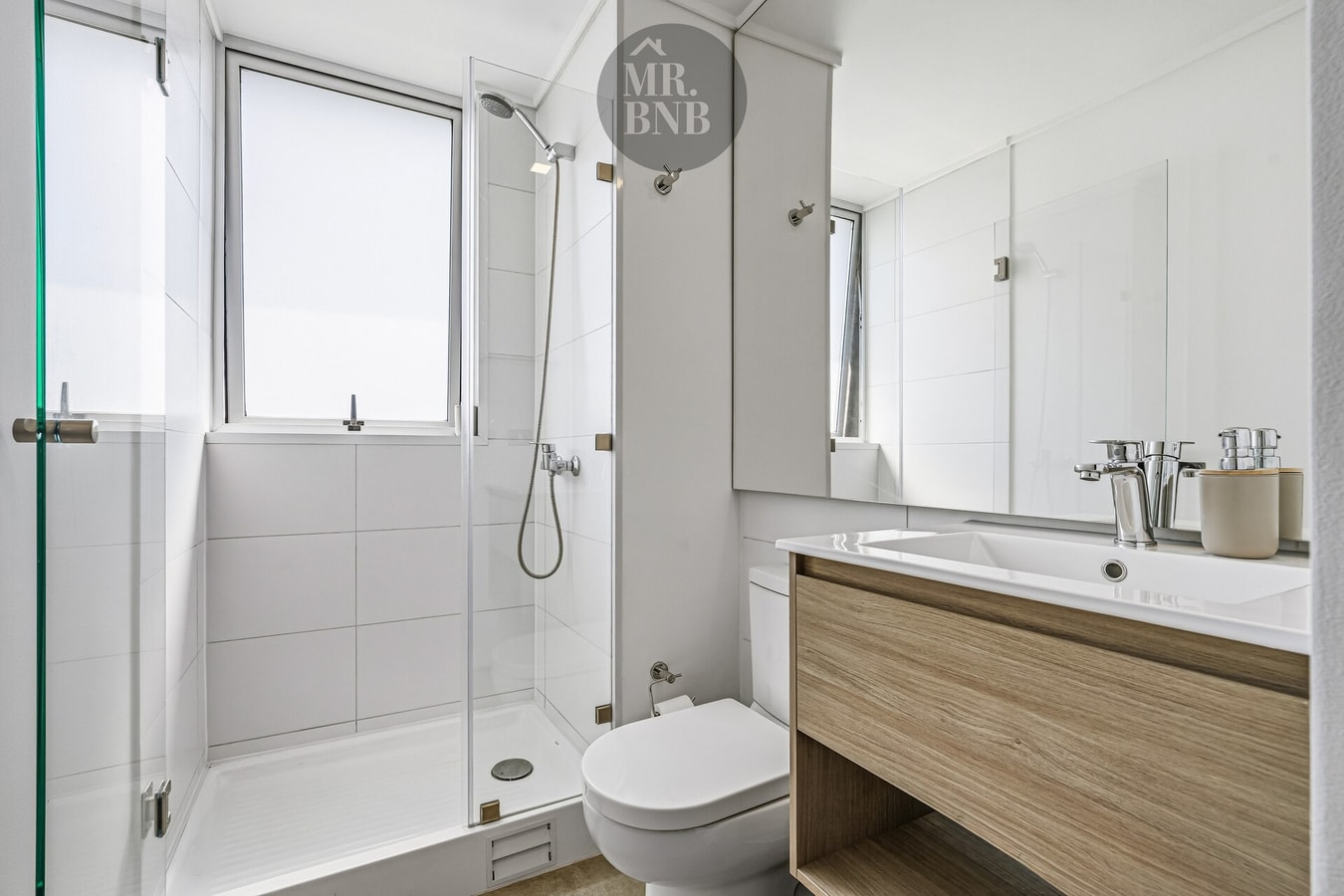Baño con ducha de vidrio, mueble vanitorio de madera y artículos de aseo