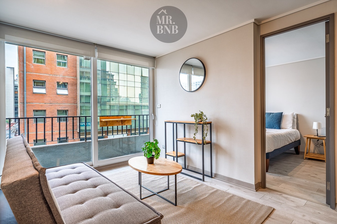 Living con sofá, ventanal amplio con vista a edificios y acceso a dormitorio.