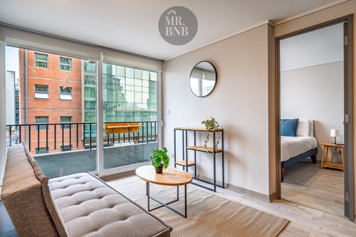 Living con sofá, ventanal amplio con vista a edificios y acceso a dormitorio.