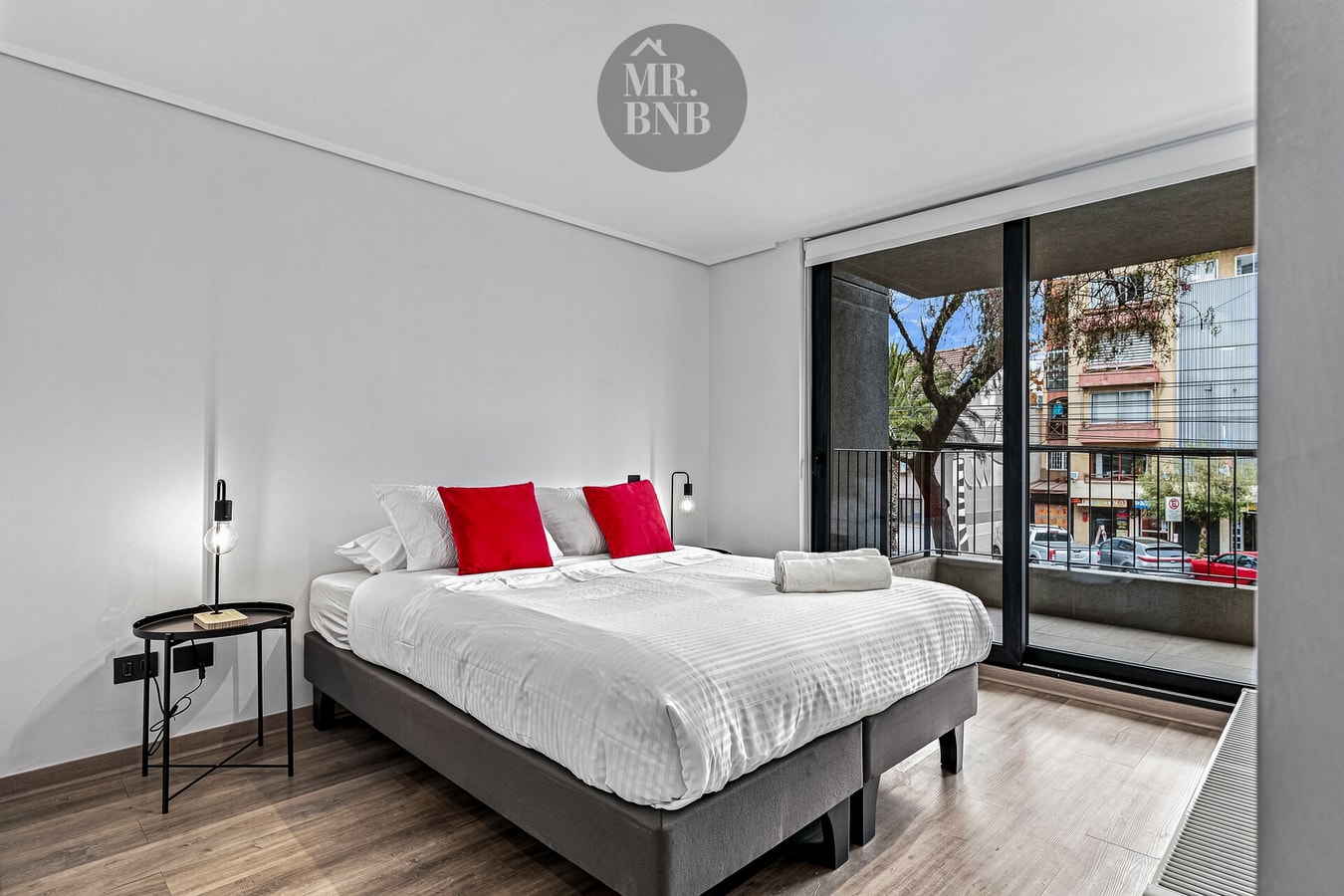 Dormitorio con cama de dos plazas, cojines rojos y ventanal con vista a la calle.
