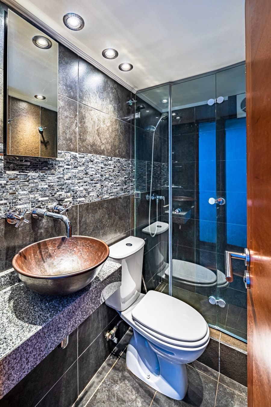 Baño con lavamanos tipo bowl de cobre, mesón oscuro, inodoro y ducha con puerta de vidrio.