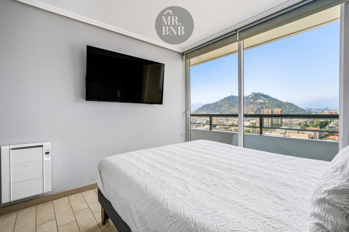 Dormitorio con cama, TV montada en la pared, radiador y ventanal con vista al cerro