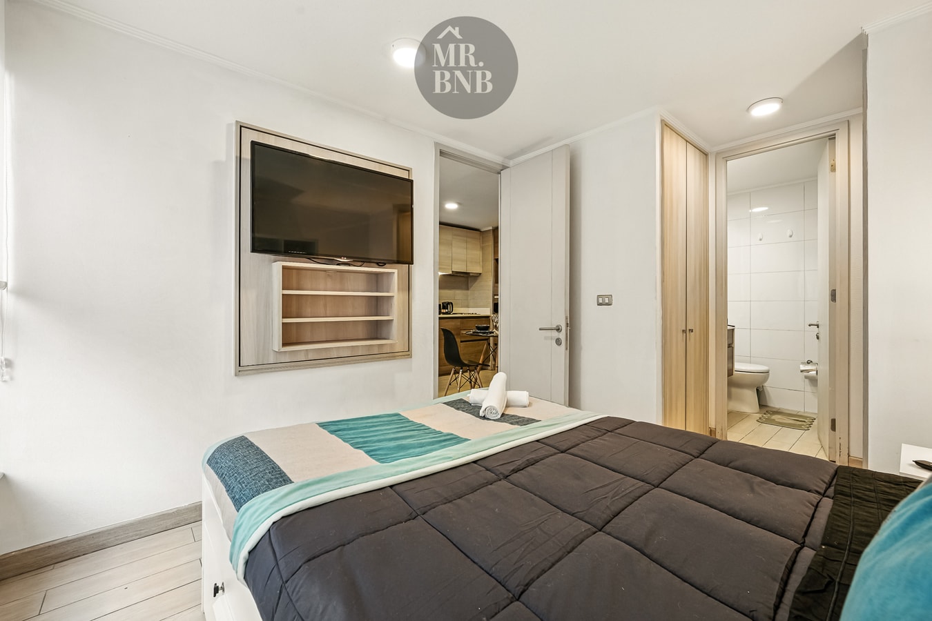 Dormitorio con cama, televisor en la pared y puerta hacia el pasillo.