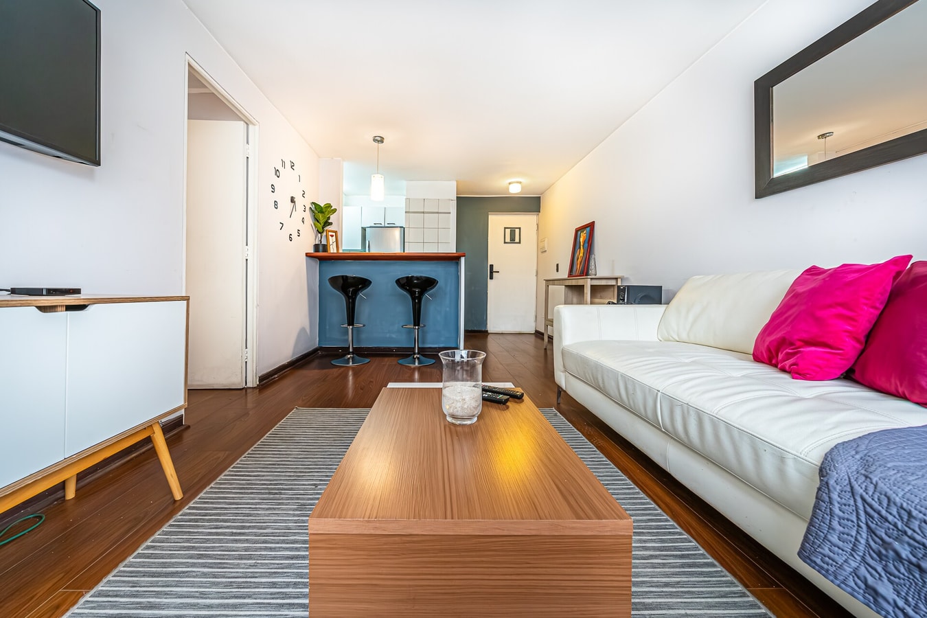 Living comedor con mesa de centro de madera, sofá blanco con cojín fucsia y barra de cocina al fondo