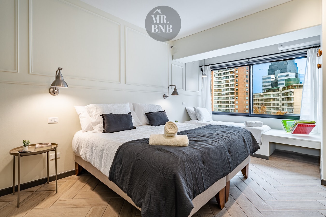 Dormitorio con cama, ventanal con vista a edificios y banca al pie de la cama