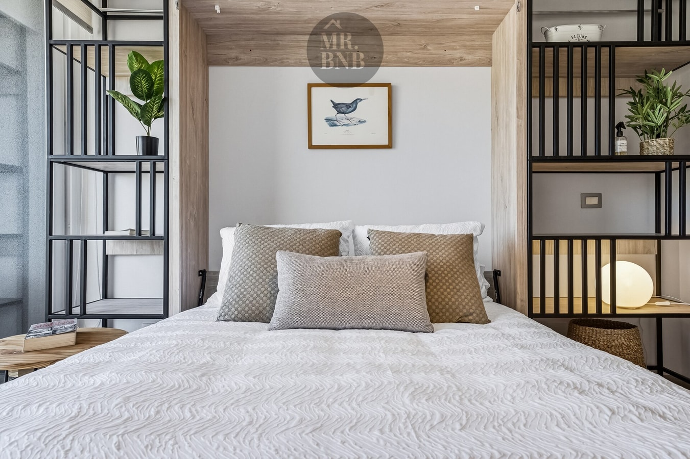 Cama con cojines junto a estantería con plantas y cuadro decorativo