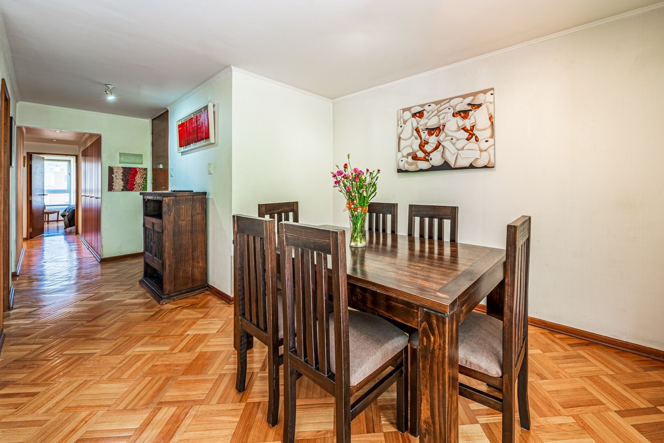 Comedor con mesa de madera para seis personas, sillas tapizadas y arreglo floral, sobre piso de parqué.