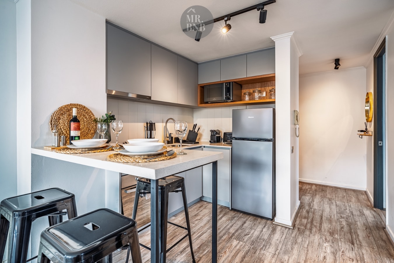 Cocina en L con muebles grises, microondas empotrado, refrigerador y barra desayunadora con banquetas