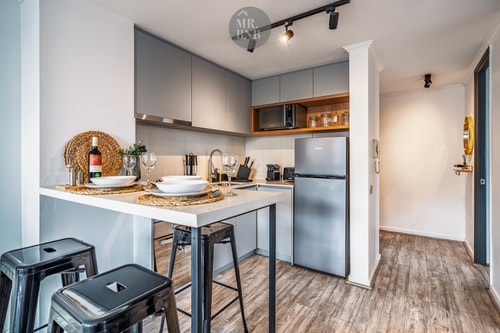 Cocina en L con muebles grises, microondas empotrado, refrigerador y barra desayunadora con banquetas