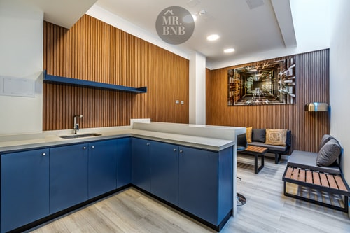 Sala de estar y cocina de un espacio común del edificio, con muebles azules y living con paneles de madera