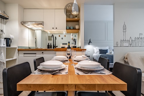 Comedor con mesa de madera puesta, sillas negras y cocina al fondo con mural de la ciudad