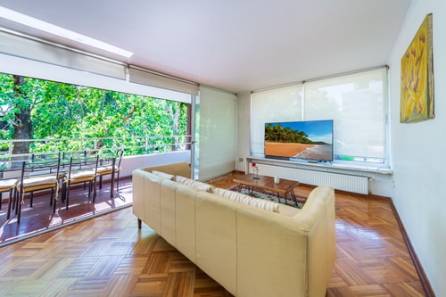 Living con sofás en L, televisor y ventanal corrido con vista a copas de árboles.