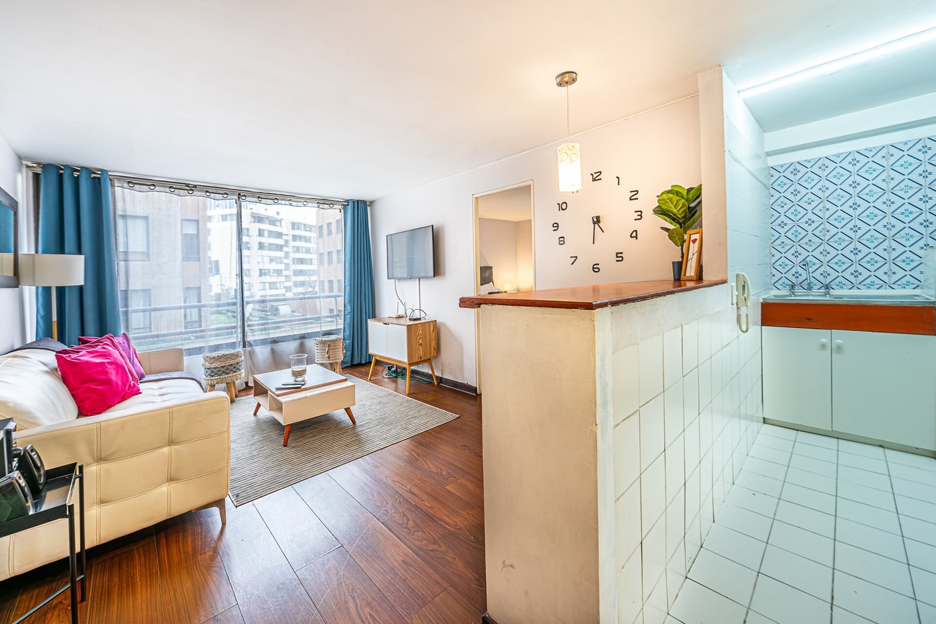 Living con barra de cocina en azulejo, televisor, reloj de pared y sofá con cojín fucsia