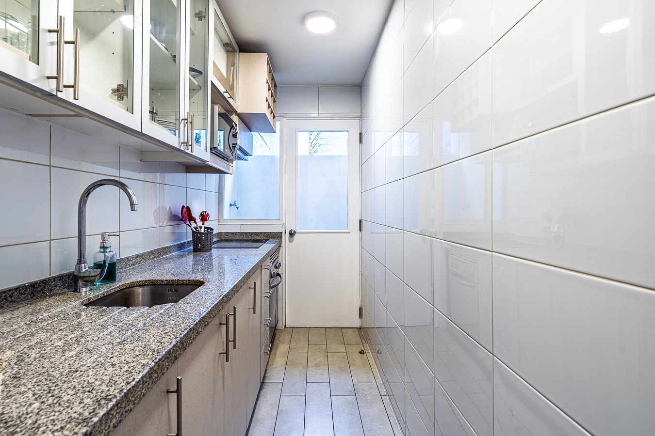 Cocina alargada con mesón de granito, muebles de vidrio y puerta hacia el exterior.