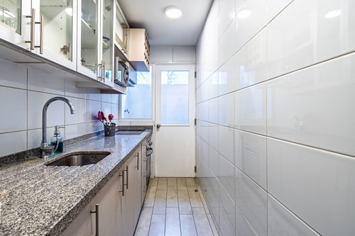 Cocina alargada con mesón de granito, muebles de vidrio y puerta hacia el exterior.