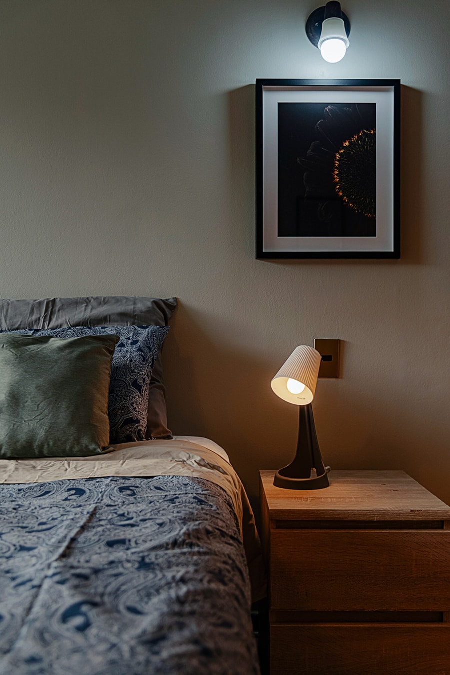 Dormitorio con cama, velador con lámpara y cuadro decorativo.