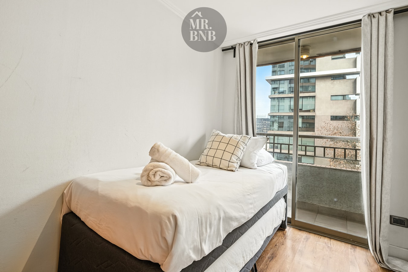 Dormitorio con cama individual junto a ventanal con cortinas y vista a edificios