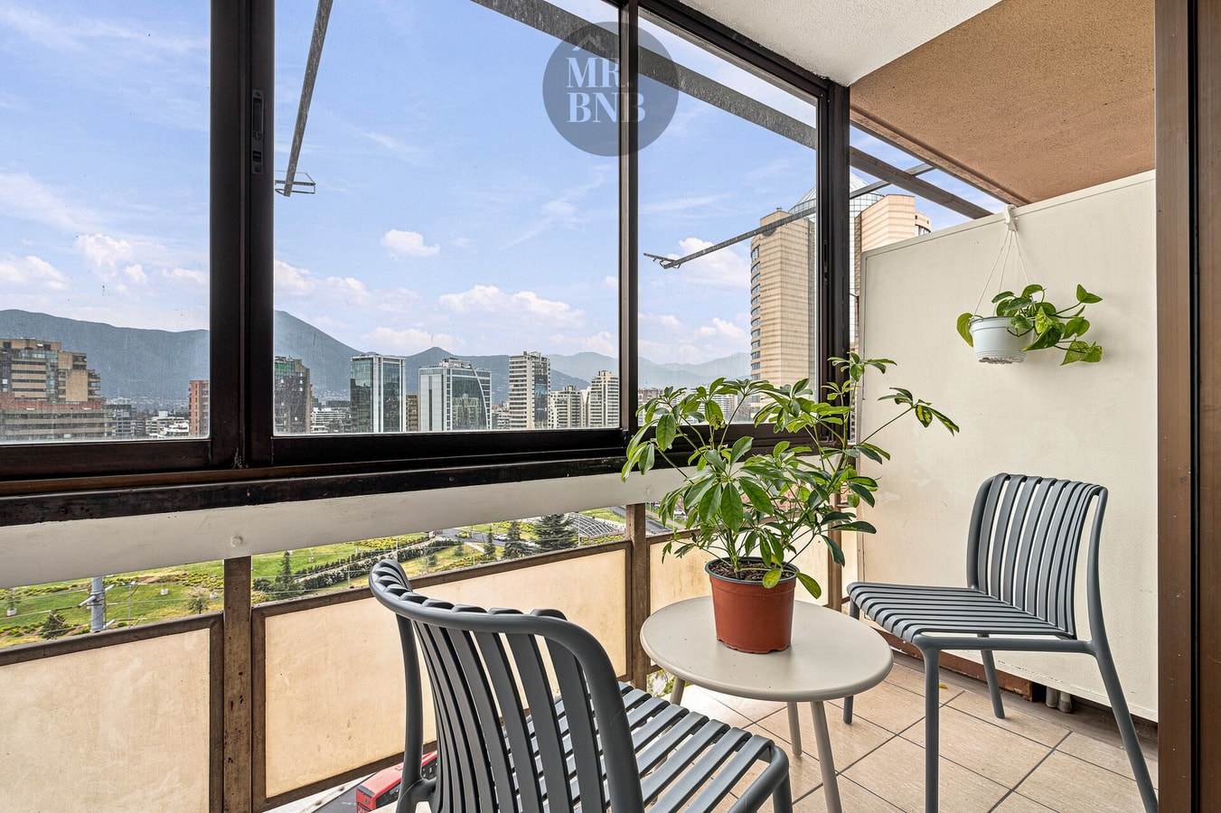 Terraza cerrada con ventanales, sillas, planta y vista despejada hacia la ciudad y la cordillera.