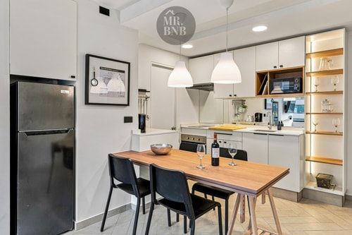 Comedor con mesa de madera, sillas negras y cocina con refrigerador al fondo.