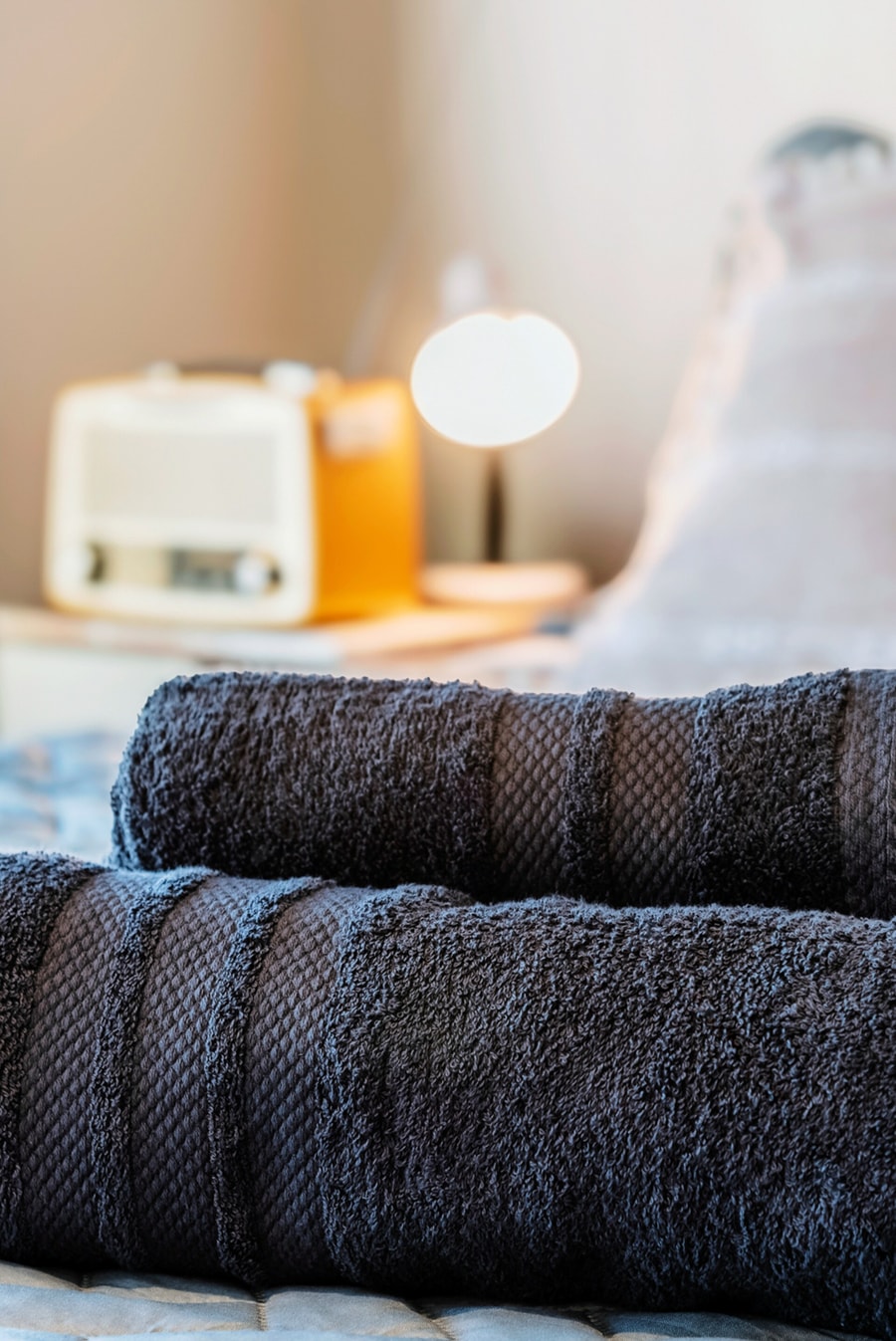 Detalle de dos toallas grises dobladas sobre la cama, con una radio retro y la lámpara encendida en el velador al fondo