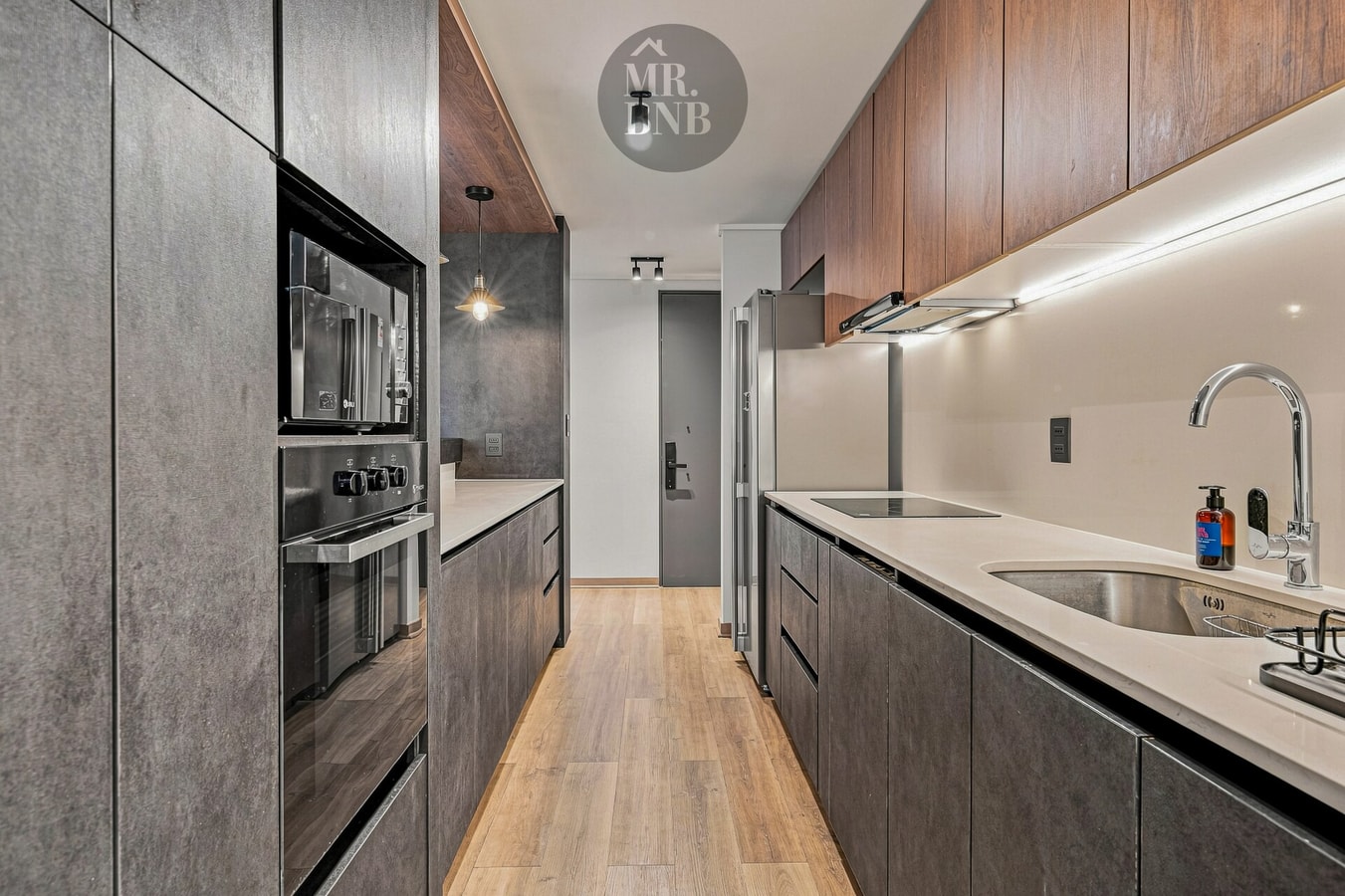 Cocina angosta con muebles oscuros, horno y microondas empotrados, y encimera clara