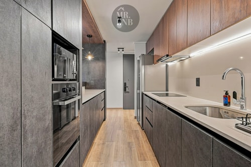 Cocina angosta con muebles oscuros, horno y microondas empotrados, y encimera clara