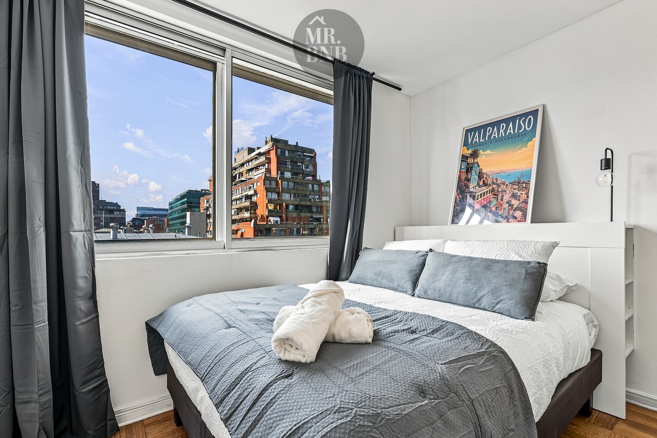 Dormitorio con cama, cuadro de Valparaíso, ventanal con cortina oscura y vista a edificios