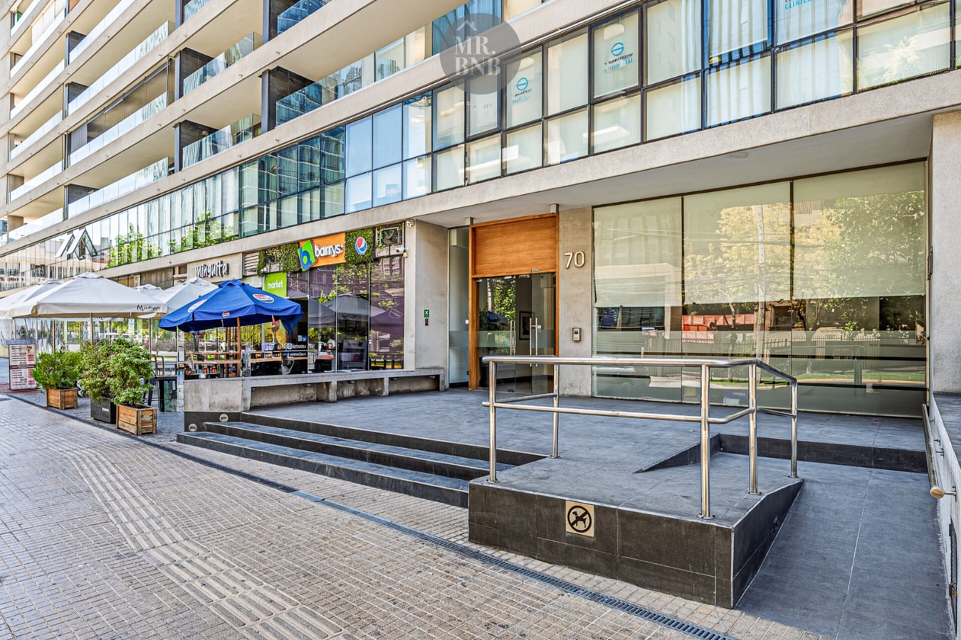 Acceso peatonal del edificio desde la vereda, con puerta de madera, el número 70, rampa con pasamanos y locales comerciales al costado