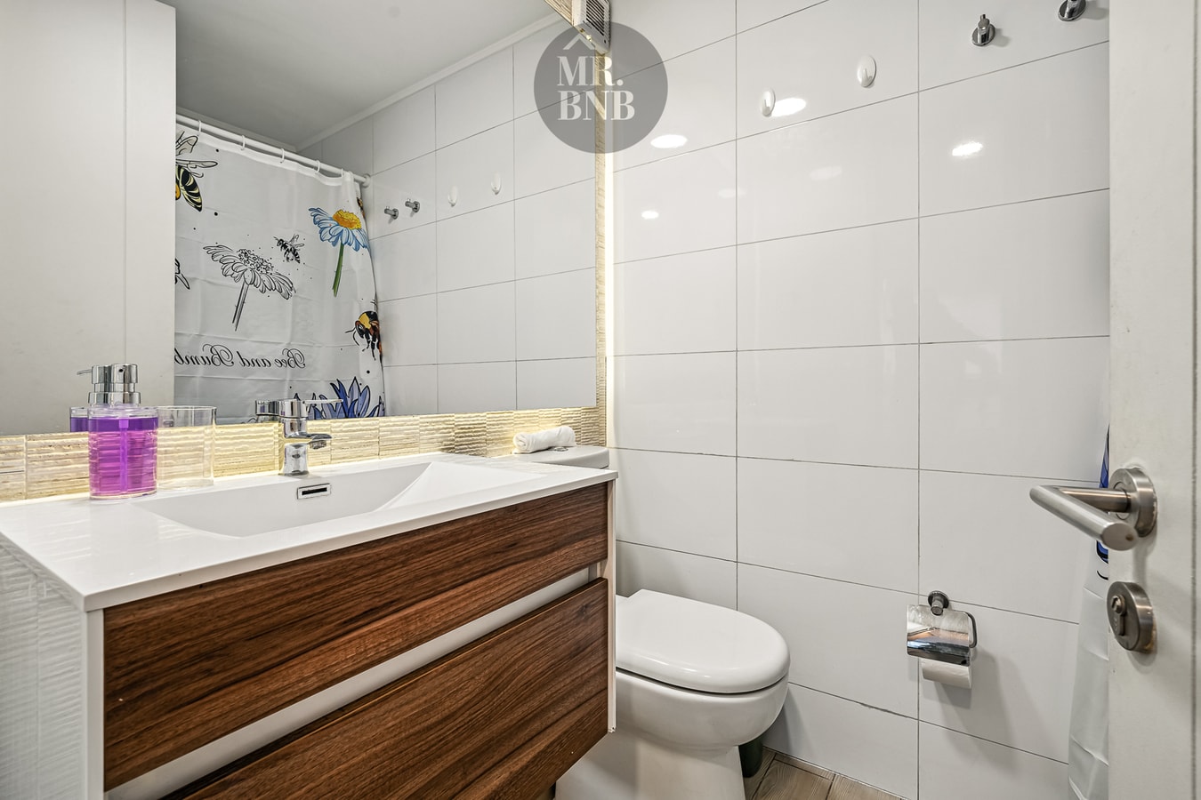 Baño con lavamanos de madera, cortina floral, inodoro y espejo.